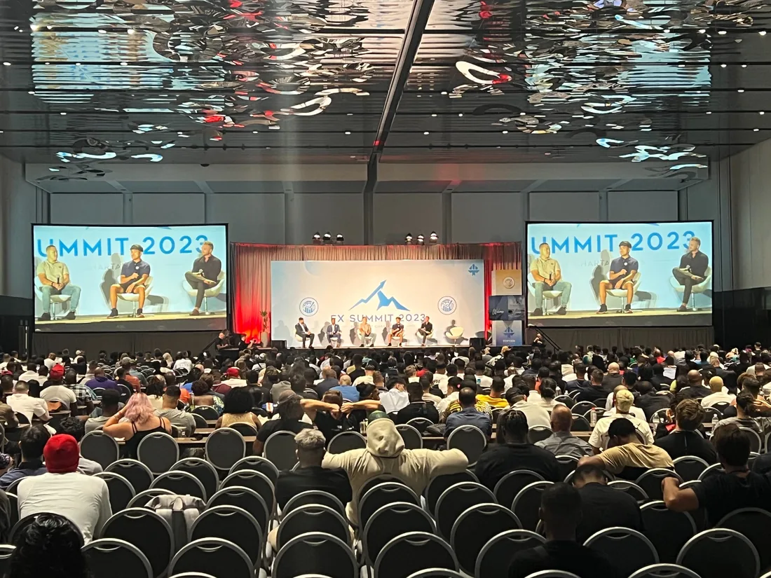 Audience facing a stage with wide projection screens at a summit in the Miami Beach Convention Center